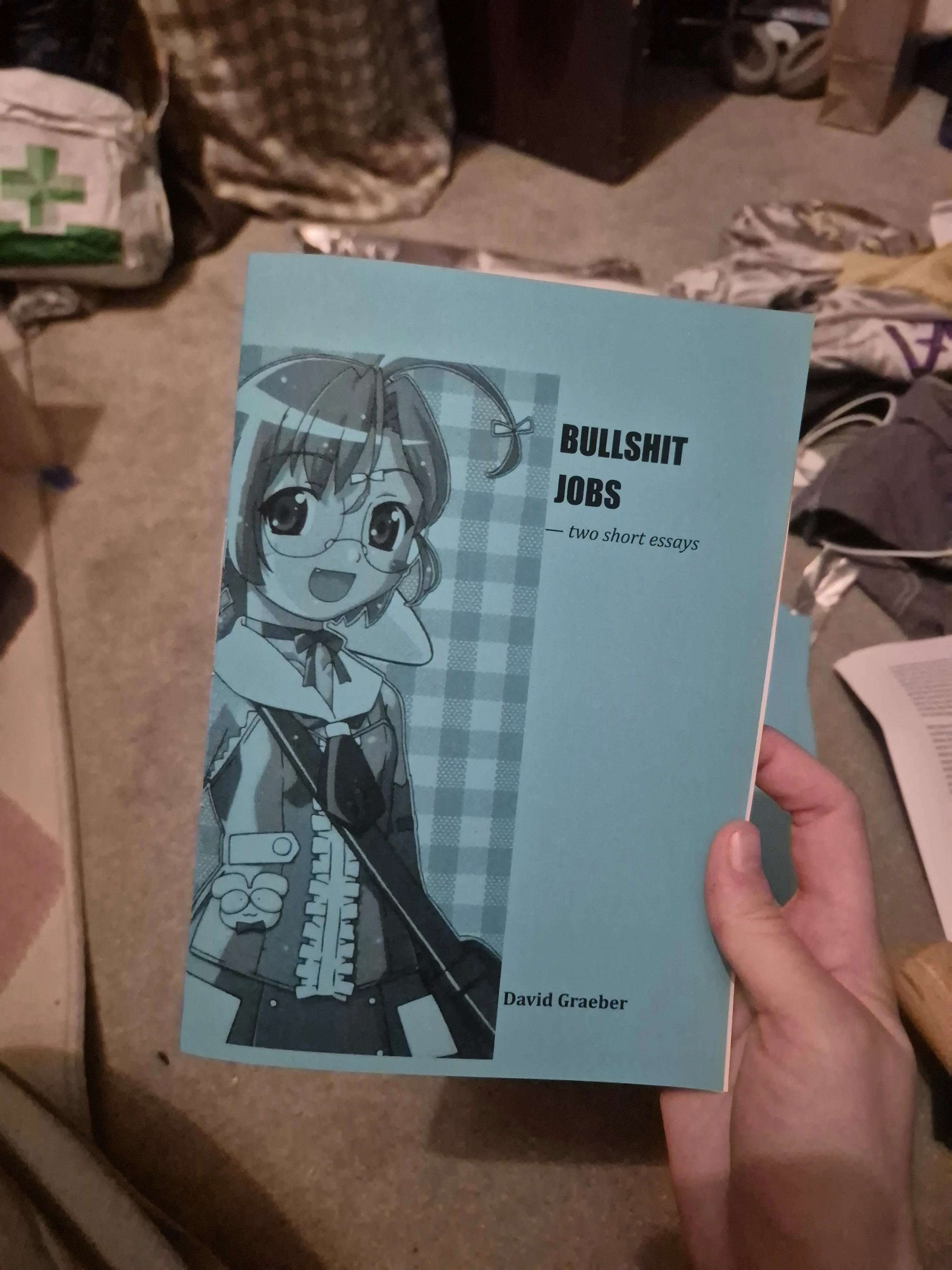 Photo of white hand holding a blue zine, titled Bullshit Jobs with an anime girl on the cover. My messy room is out of focus in the background.